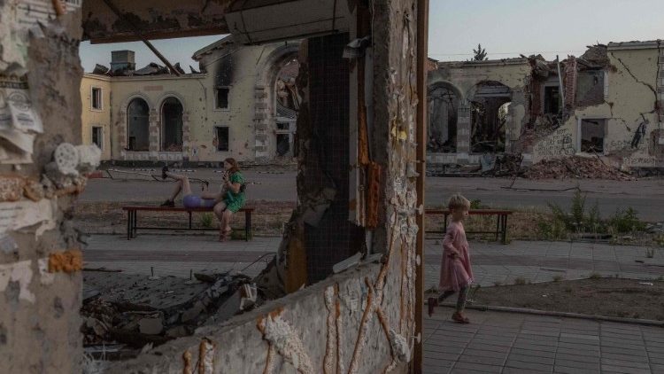 Uma criança brinca enquanto mulheres descansam em um banco em frente a prédios residenciais destruídos por bombardeios em Kostyantynivka, região leste de Donetsk, em 22 de junho de 2024. (Foto de Roman PILIPEY/AFP)