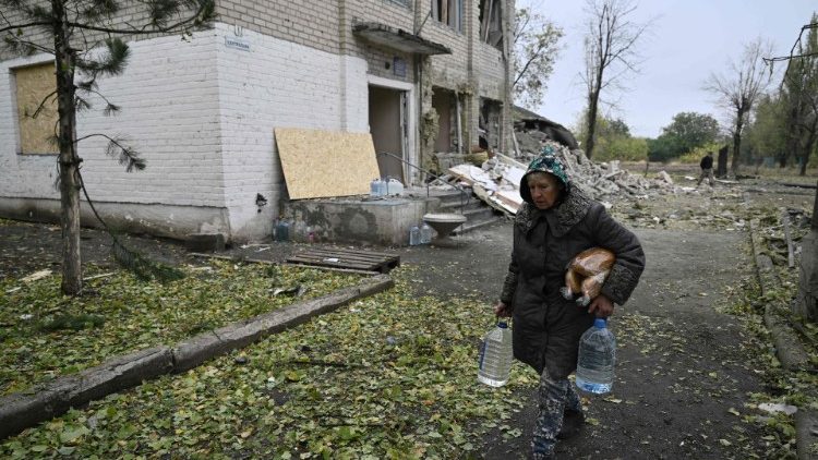 Uma moradora local carrega pão e água enquanto passa por um prédio administrativo local, destruído após o ataque russo na vila de Shevchenko, perto da cidade de Pokrovsk, região de Donetsk, em 20 de outubro de 2024, em meio à invasão russa da Ucrânia (Foto de Genya SAVILOV / AFP)
