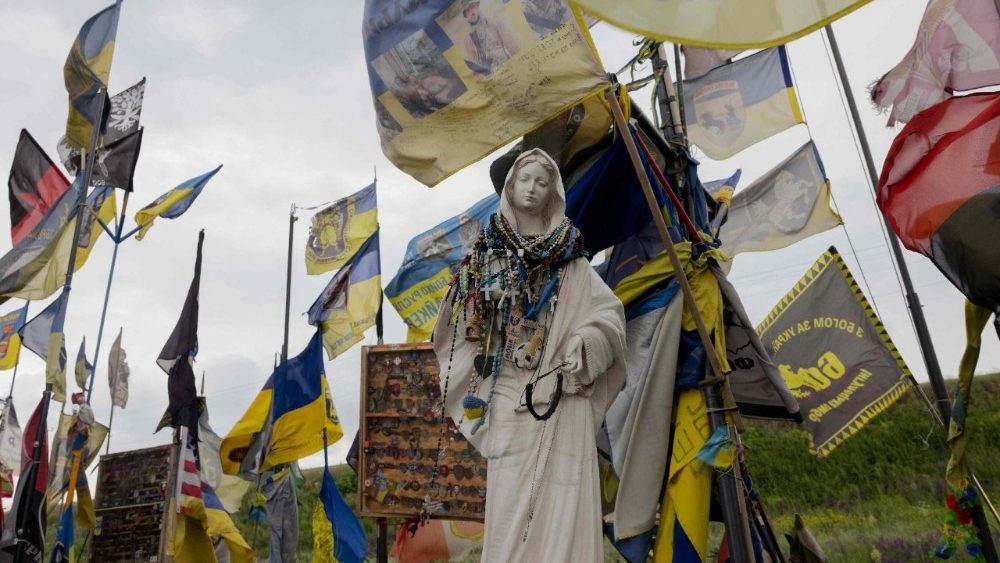 Esta fotografia mostra uma estátua de Nossa Senhora entre as bandeiras das brigadas ucranianas em frente à placa de entrada da região de Donetsk, protegida por uma rede antidrone, em 19 de junho de 2025, em meio à invasão russa da Ucrânia. (Foto de Tetiana DZHAFAROVA / AFP)   (AFP or licensors)