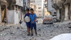 Crianças palestinas segurando recipientes de água caminham sobre os escombros de um prédio alvo de um ataque israelense no bairro de Sheikh Radwan, na Cidade de Gaza, no centro da Faixa de Gaza, em 6 de julho de 2025.. (Foto de Eyad BABA / AFP)
