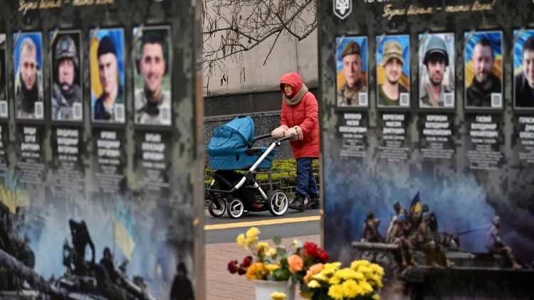 Uma mulher com um carrinho de beb&ecirc; caminha ao lado de retratos ao longo da Alameda dos Her&oacute;is Ca&iacute;dos, em um distrito de Kiev, em 7 de dezembro de 2025, em meio &agrave; invas&atilde;o russa na Ucr&acirc;nia. (Foto de Sergei SUPINSKY / AFP)