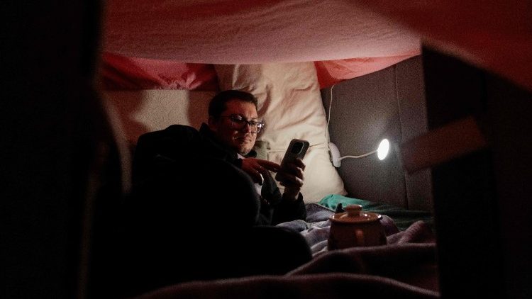 Pavlo, morador local e marido de Evgenia, senta-se sob uma tenda improvisada com travesseiros e cobertores dentro de casa para se aquecer durante um apag&atilde;o em Kiev, na noite de 13 de janeiro de 2026, em meio &agrave; invas&atilde;o russa da Ucr&acirc;nia. (Foto de Tetiana DZHAFAROVA / AFP)
