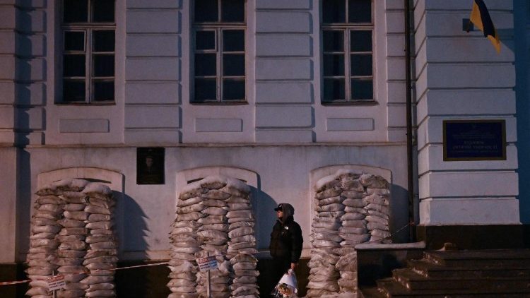 Um homem segurando flores est&aacute; ao lado de um pr&eacute;dio com as janelas protegidas por sacos de areia contra poss&iacute;veis danos causados ​​por ataques a&eacute;reos em Kiev, em 29 de janeiro de 2026, em meio &agrave; invas&atilde;o russa da Ucr&acirc;nia. (Foto de Sergei Gapon / AFP)