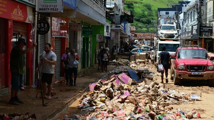 Pessoas caminham por uma rua lamacenta passando por produtos descartados no distrito de Ub&aacute;, em Minas Gerais.