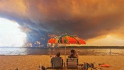 Beach goers sit on the beach of Moutchic in Lacanau, southwestern France on July 24, 2026, as clouds of smoke rise into the sky from a wildfire.