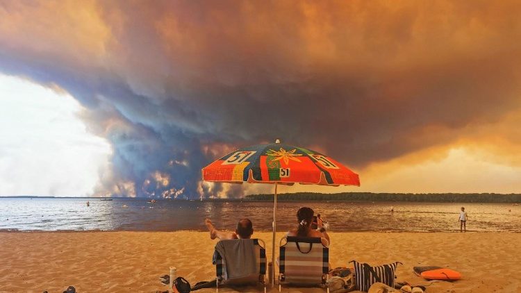 Beach goers sit on the beach of Moutchic in Lacanau, southwestern France on July 24, 2026, as clouds of smoke rise into the sky from a wildfire.